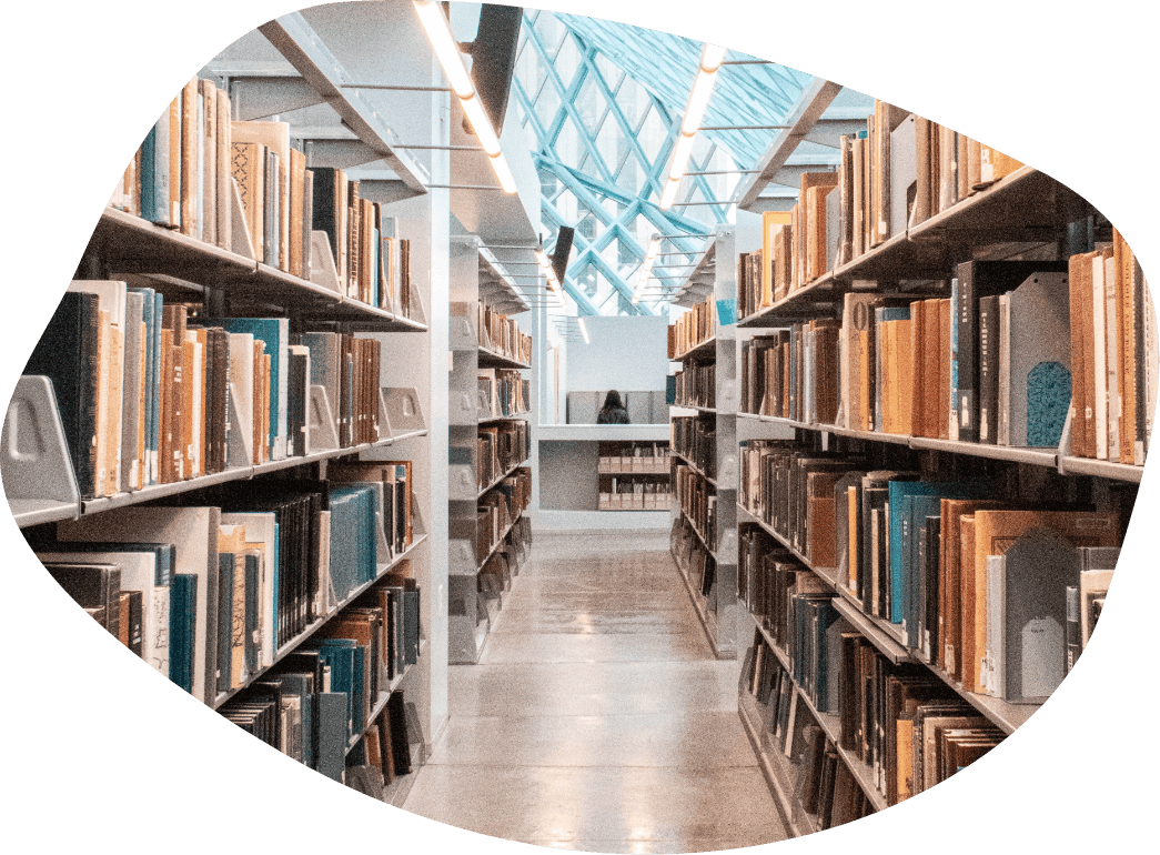 A brightly lit library aisle with shelves full of books on both sides. The aisle leads to a well-lit area with large windows in the background, allowing natural light to flood the space. The modern design features a high, angled ceiling, making it an ideal place for education and recruitment activities.