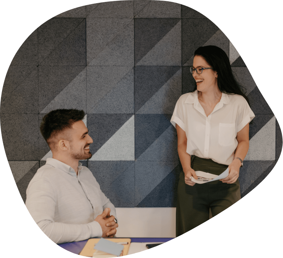 A man seated at a desk and a woman standing next to him are engaged in a cheerful conversation. The man has a notepad and sticky notes in front of him, while the woman holds papers. They are in an office with a geometric patterned wall, likely discussing how to get hired successfully.