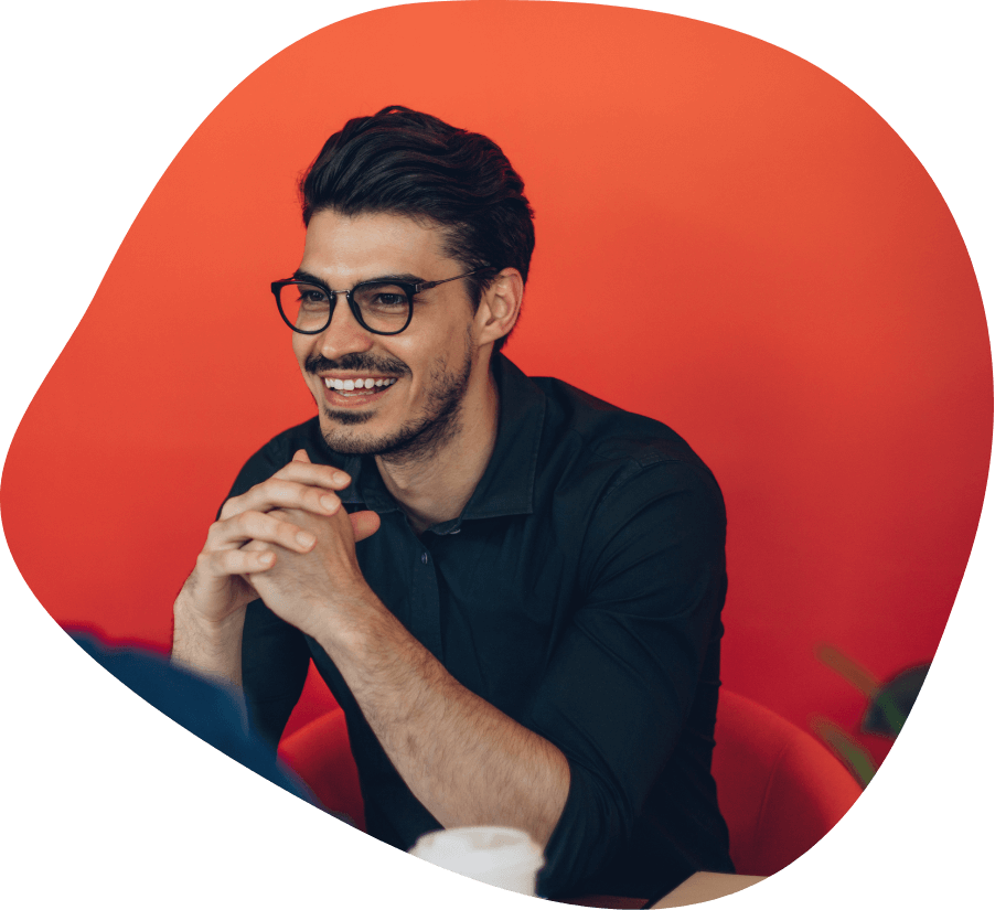 Person with short hair and glasses smiling, wearing a dark shirt, sitting against a bright red background, ready to get hired.
