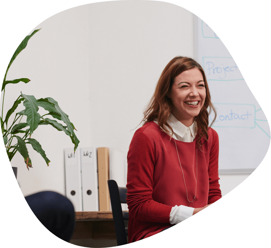 A woman with shoulder-length brown hair is sitting and smiling in an office setting, possibly engaged in office support. She is wearing a red sweater over a white collared shirt. Behind her are whiteboards with diagrams and two binders on a shelf. A green plant is visible to her left.