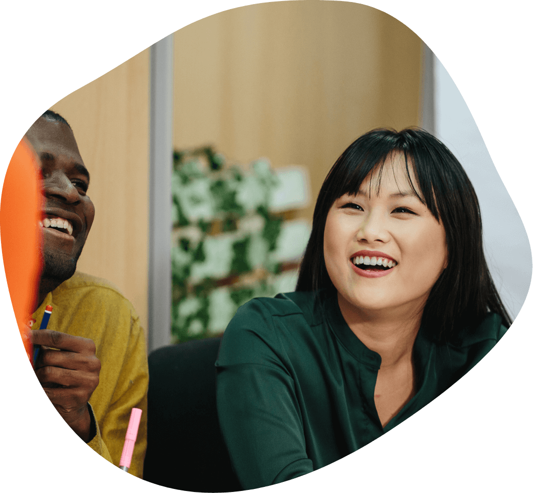 Two people are sitting together and smiling. The person on the left is wearing a yellow shirt. The person on the right, with long dark hair and wearing a green top, is laughing. They are indoors, possibly discussing education recruitment, with a plant and wooden wall visible in the background.