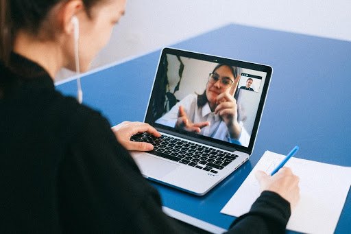 A woman wearing earbuds is seated at a blue table, using a laptop for a video interview with a person depicted on the screen. She is holding a pen and writing on a notepad placed beside the laptop.