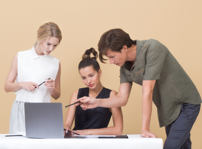 Three people are gathered around a laptop, fostering a feedback culture. Two women are sitting, and the man is leaning over, pointing at the screen. The background is a solid beige color. The woman on the left is holding a pen, while the woman in the center is focused on the laptop screen.