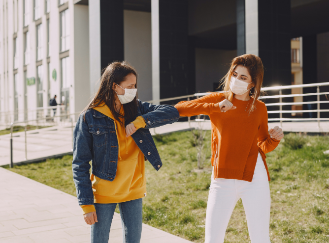 Two women wearing face masks greet each other by touching elbows outdoors, embracing the engaging culture. One woman wears a denim jacket over a yellow hoodie and jeans, while the other wears an orange sweater and white pants. They stand on a paved pathway with a modern building in the background, reminiscent of Rich Roberts' architectural style.