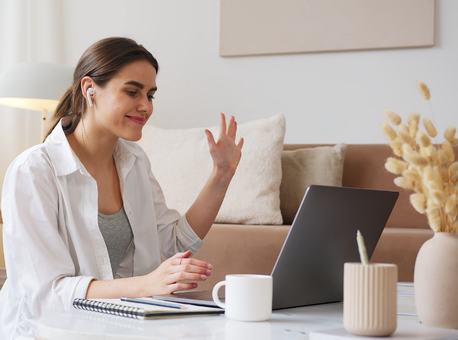 A woman with brown hair wearing a white shirt waves at her laptop screen while engaging in a video call. She is sitting at a table with a white mug, a notebook, a pen holder, and dried flowers. A beige couch and cushion are in the background as she manages teams remotely.