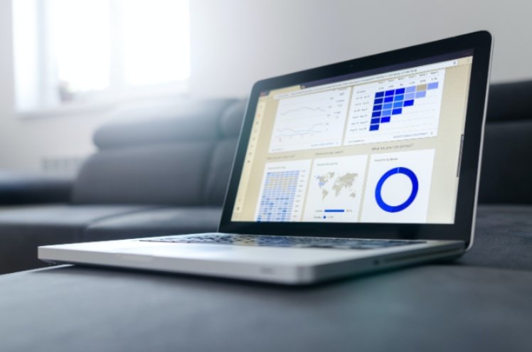 A laptop displaying various data analytics and charts, including a bar graph, a world map, and a circular chart, rests on a dark cushion in a well-lit office support recruitment room with windows in the background.