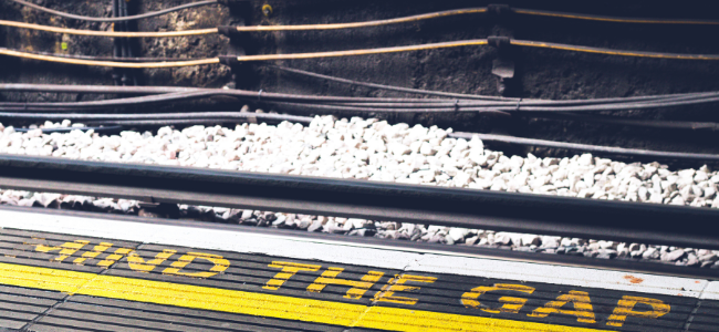 A train platform with a yellow line and the words "MIND THE GAP" painted on the edge. Beyond the edge, white gravel lies alongside tracks and a dark, textured wall in the background, reminiscent of 2021's bustling scenes as commuters awaited legal updates on employment laws.