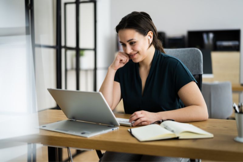 A woman sitting at a desk is smiling while looking at a laptop screen, her dark green top complementing the star-patterned earpiece in her right ear. An open notebook, a pen, and a cup of coffee are on the desk. The background shows an office setting.