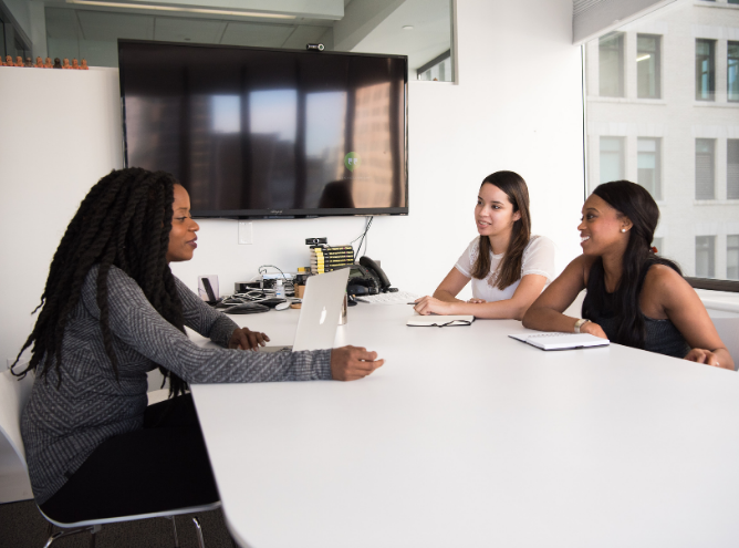 Three women sit at a white conference table in an office. One woman, on the left, works on a laptop while discussing inclusive hiring techniques. The other two women listen and take notes. There's a large, wall-mounted screen and windows with an exterior building view in the background.