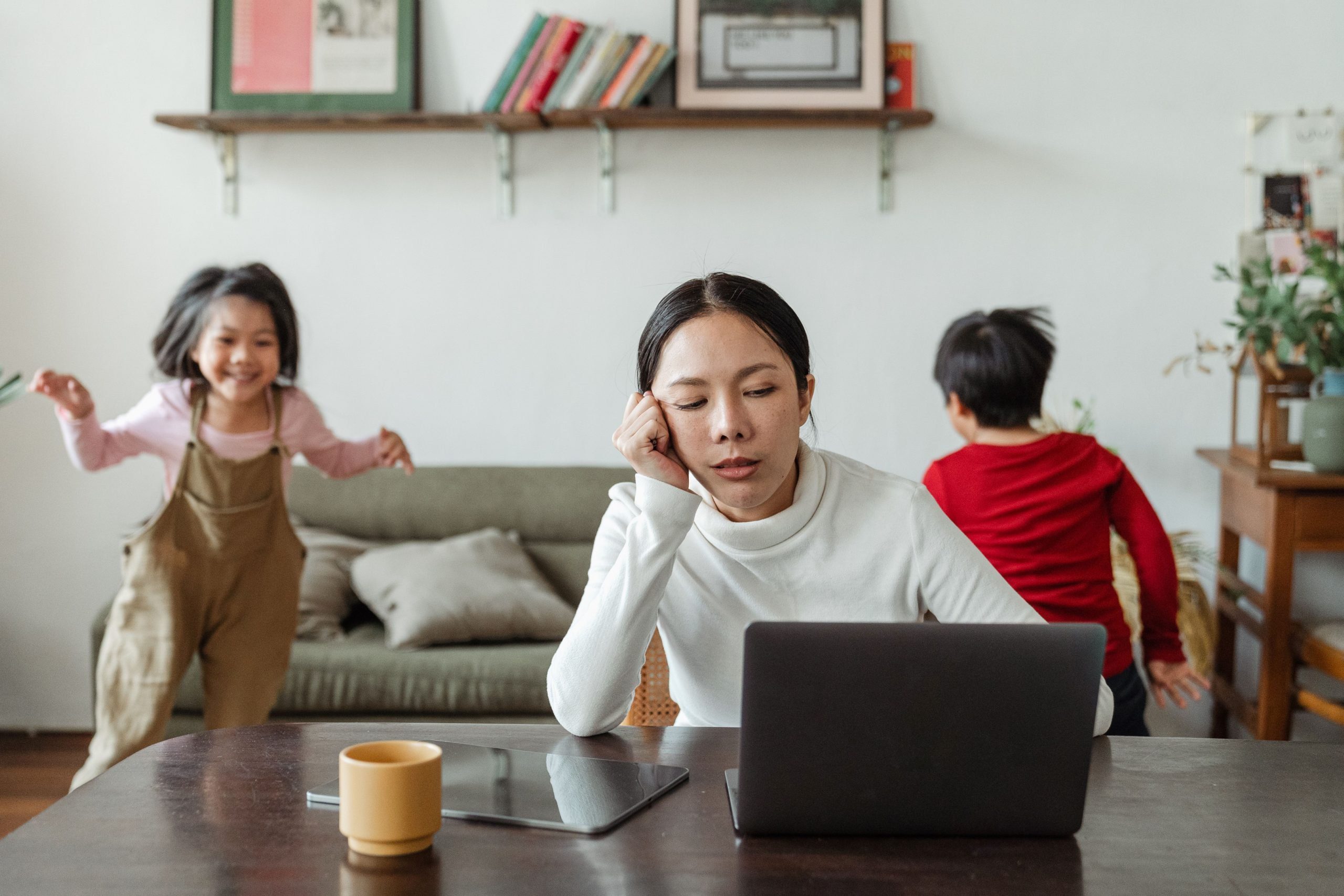 A woman appears tired while working on a laptop at a table. A coffee cup and tablet are beside her, symbols of her effort to stay productive. In the background, two children play energetically in an inclusive living room with a sofa, bookshelf, and framed pictures on the wall.