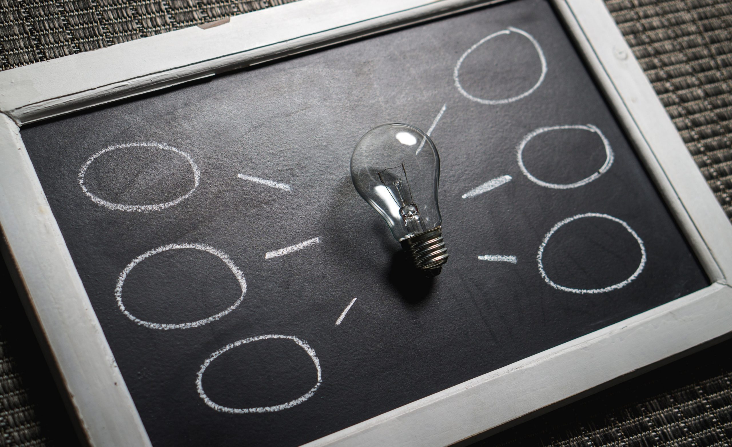 A clear light bulb sits in the center of a small chalkboard. Surrounding it are six empty circles drawn with chalk, each connected to the bulb by lines, suggesting a productive return to work concept. The chalkboard's frame is wooden.