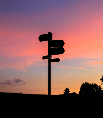 A silhouette of a signpost with multiple directional arrows stands against a colorful sunset sky, featuring hues of purple, pink, orange, and yellow. Trees and a horizon are visible in the distance, much like the evolving market trends of 2022 guiding us through new directions.