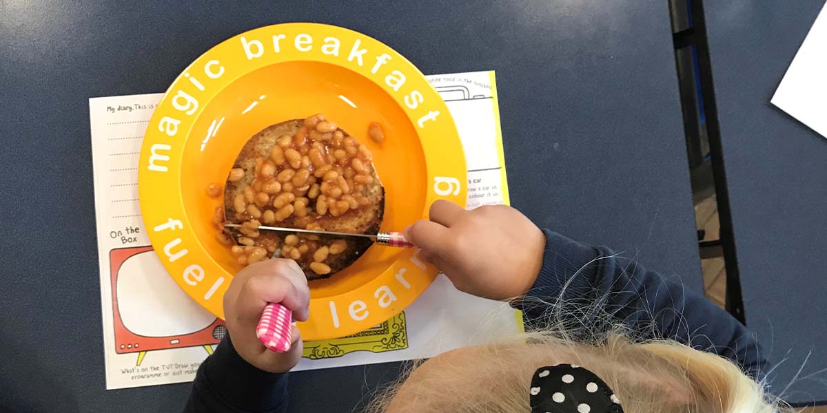 Magic Breakfast A child with a black polka dot bow in their hair is using a fork and knife to enjoy baked beans on toast from a yellow plate that reads, "magic breakfast" and "fuel for learning." Seated at a dark table with papers underneath the plate, they appear focused on their meal.
