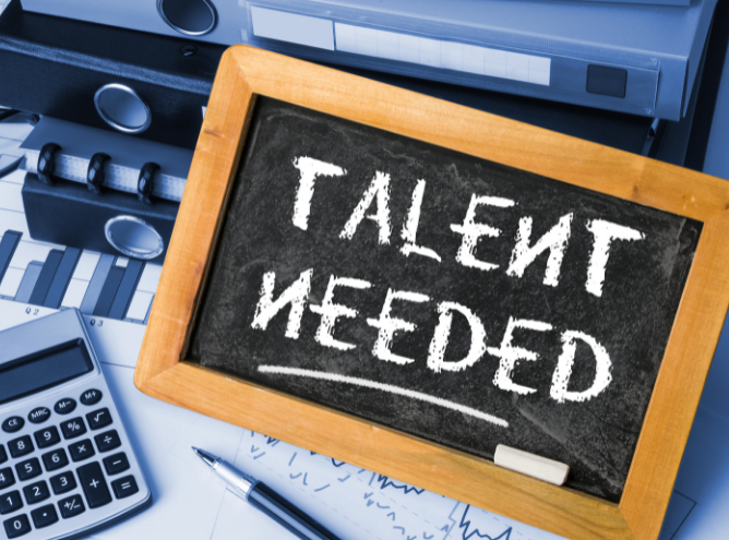 A small chalkboard with the words "TALENT NEEDED" written in white chalk is surrounded by office supplies, including a calculator, binders, a pen holder, and a financial graph; it's clear that securing the best talent is crucial in this bustling office.