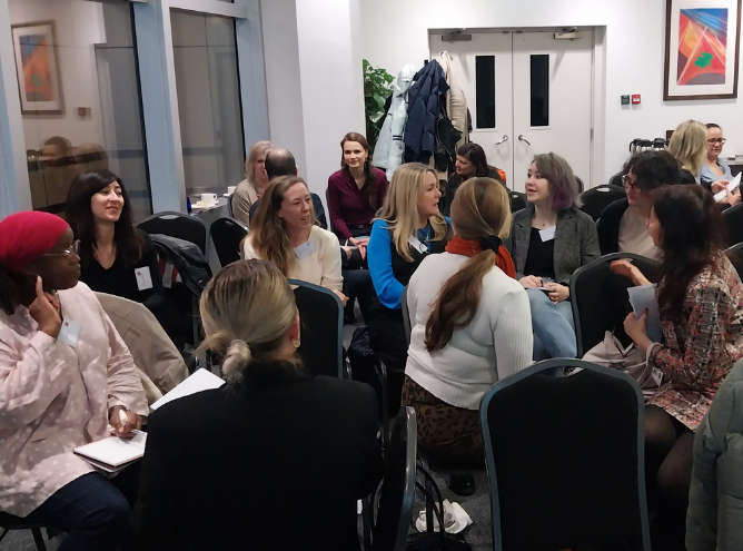 In March 2023, a group of women are sitting and standing in a room, engaged in lively conversation. Some are facing each other in small clusters, holding notebooks or papers. Coats and bags hang on hooks by the doors in the background. The room has white walls and colorful artwork from the Signature Leadership Breakfast event.