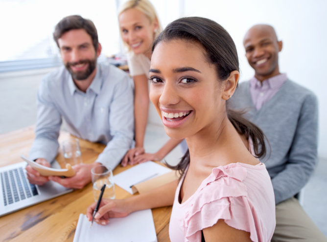 A group of four diverse individuals is gathered around a wooden table, smiling and looking at the camera. A laptop, papers, and a glass of water are visible on the table. One person in the foreground is writing down amazing strategies on a notepad.