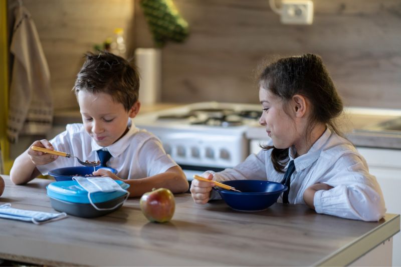 Two children in school uniforms sit at a kitchen table eating from blue bowls. A packed lunch container and an apple are on the table. The kitchen in the background has a stove and various utensils, suggesting a busy morning routine for parents juggling jobs for graduates. The children appear to be enjoying their meal.