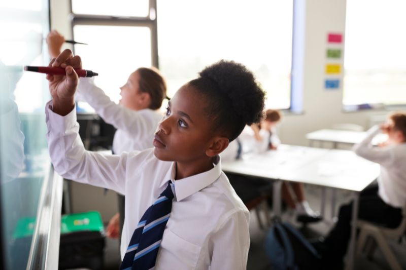 A student in a school uniform writes on a whiteboard with a pen while several other students work at desks in the background. The classroom, adorned with education-related posters and bright natural light streaming through the windows, buzzes with future achievers considering jobs for graduates.