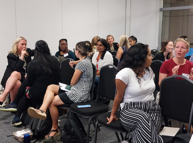 In June 2023, a group of people is sitting on black chairs in a conference room, engaged in conversations about leadership. Some are in pairs while others chat in small groups. There's a mix of men and women, casually dressed. A few bags and personal items are placed on the floor beside them.