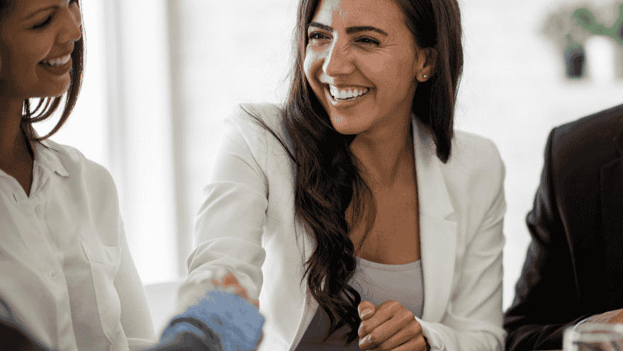 A woman in a white blazer is smiling and shaking hands with another person, perhaps discussing a temp work opportunity. She is seated beside two other people in a professional setting discussing client success stories.