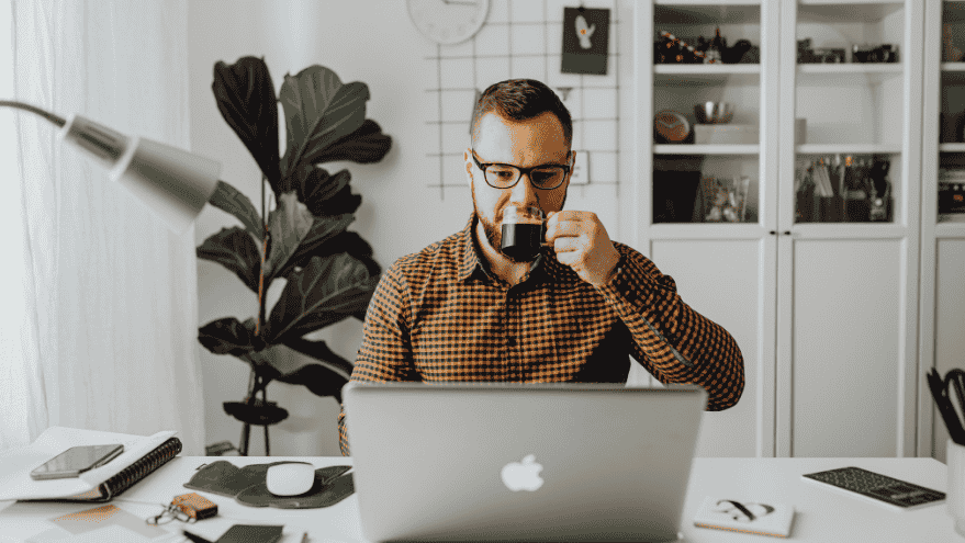 A man with glasses sits at a desk, on his laptop on a job search overqualified and drinking from a black mug. The modern workspace features a plant, notebooks, and shelves in the background.